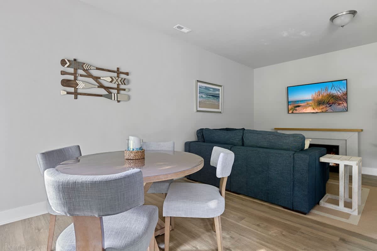 Dining area featuring a round wooden table with a glass top and four upholstered chairs, positioned next to the living room with a view of the wall-mounted TV.