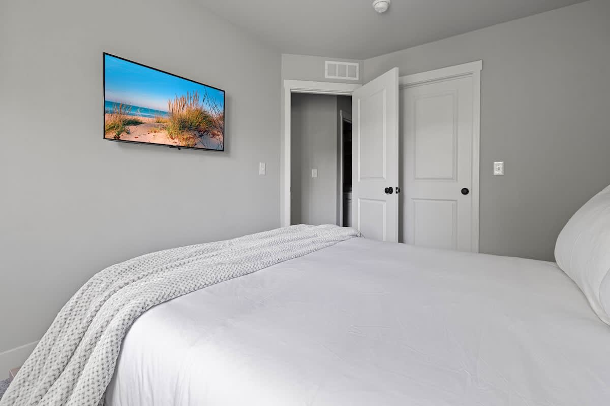 A bright bedroom with light gray walls features a large bed with white bedding and a gray throw blanket, and a wall-mounted TV displaying a vibrant beach scene. A white door with black hardware is visible in the background.