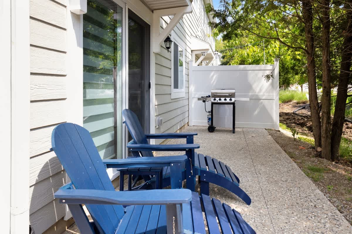 A concrete patio area features two blue Adirondack chairs and a stainless steel BBQ grill, set against the light-colored siding of the house and surrounded by trees and a white fence.