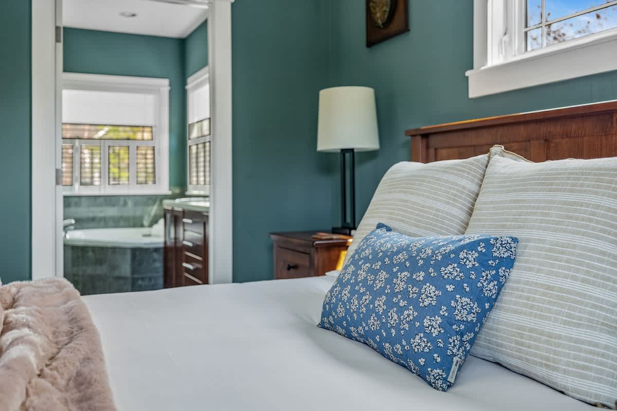 A close-up of a bed with white linens and a blue floral accent pillow, showing a wooden headboard and a nightstand with a lamp. In the background, an open doorway reveals an ensuite bathroom with a bathtub.