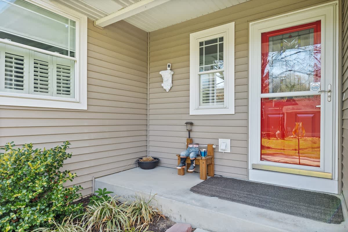 A welcoming front porch with light brown siding, a bright red storm door, and white trim, featuring a small wooden bench and lush green bushes.