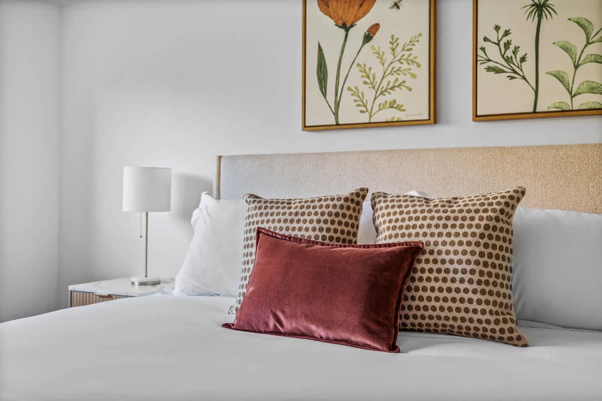 Close-up of a king-sized bed with a light upholstered headboard, white bedding, patterned pillows, and a deep red accent pillow, with botanical artwork on the wall.