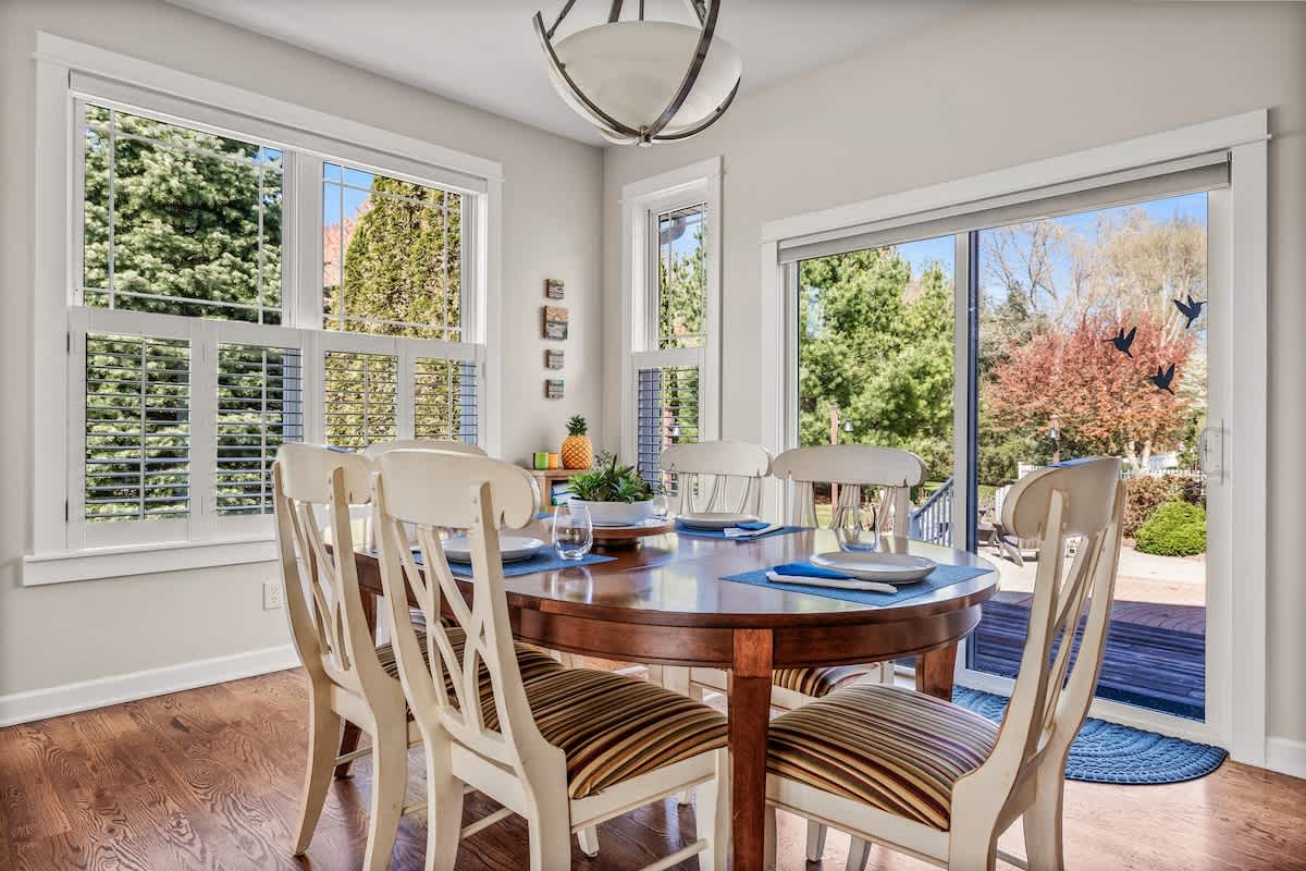 Bright dining room with a round wooden table set with blue placemats and white chairs with striped cushions. Large windows and a sliding glass door offer views of the lush outdoor landscape.