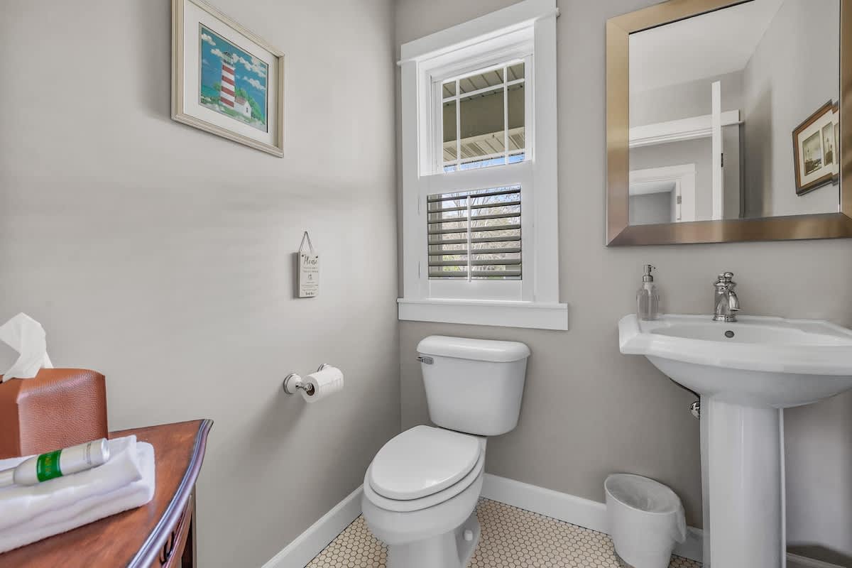 A clean half-bathroom with light gray walls, a white pedestal sink, a toilet, a small window with blinds, and a framed lighthouse picture on the wall.