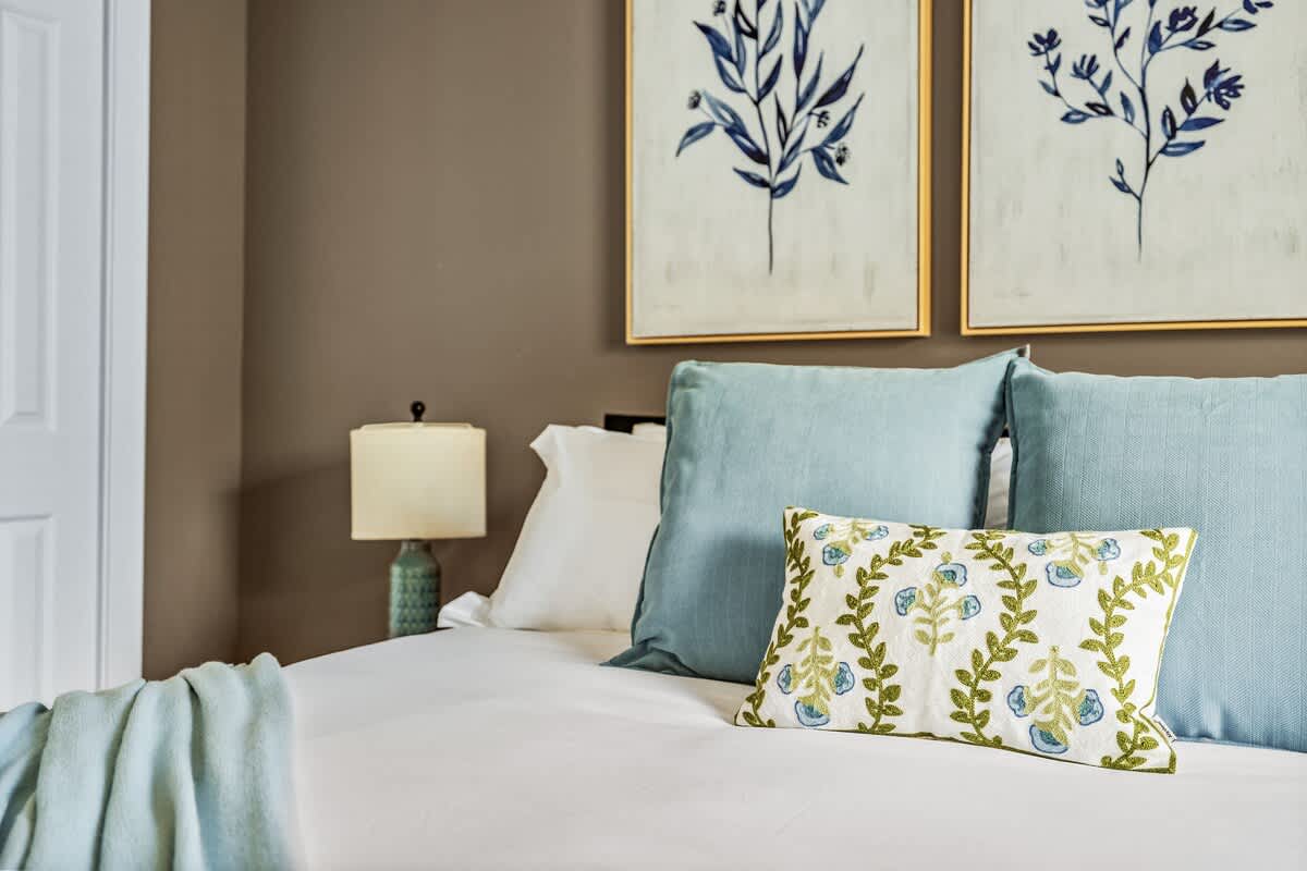 Close-up of a king-sized bed with crisp white bedding, light blue pillows, and a decorative white and green floral accent pillow, set against a warm brown wall with botanical art.