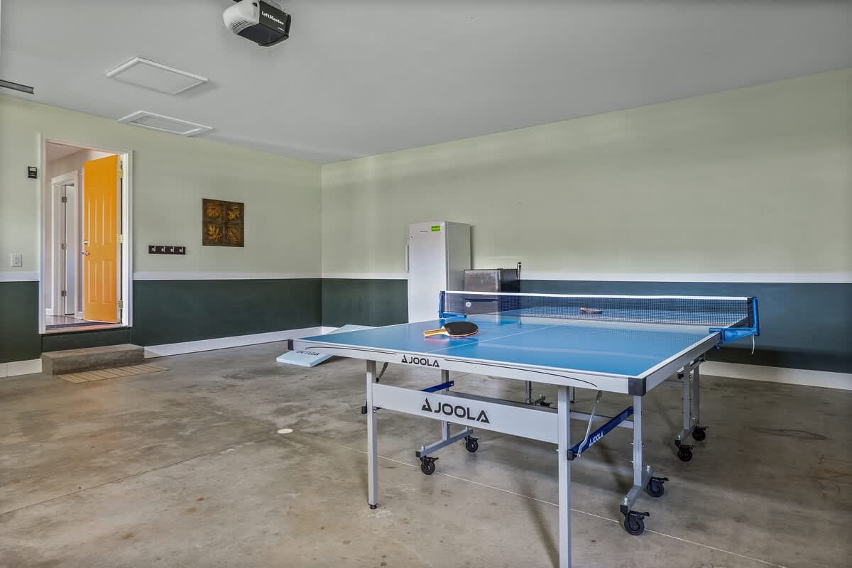 Wide view of a garage game room featuring a blue Joola ping pong table, a white refrigerator, and an orange interior door, with concrete flooring.