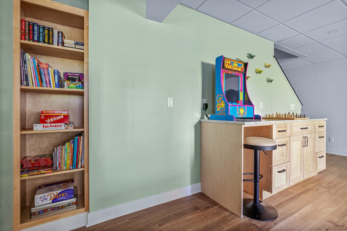 Dedicated game corner featuring a retro arcade machine, light wood built-in cabinets with a chess set, a bar stool, and a tall bookshelf filled with books and board games.