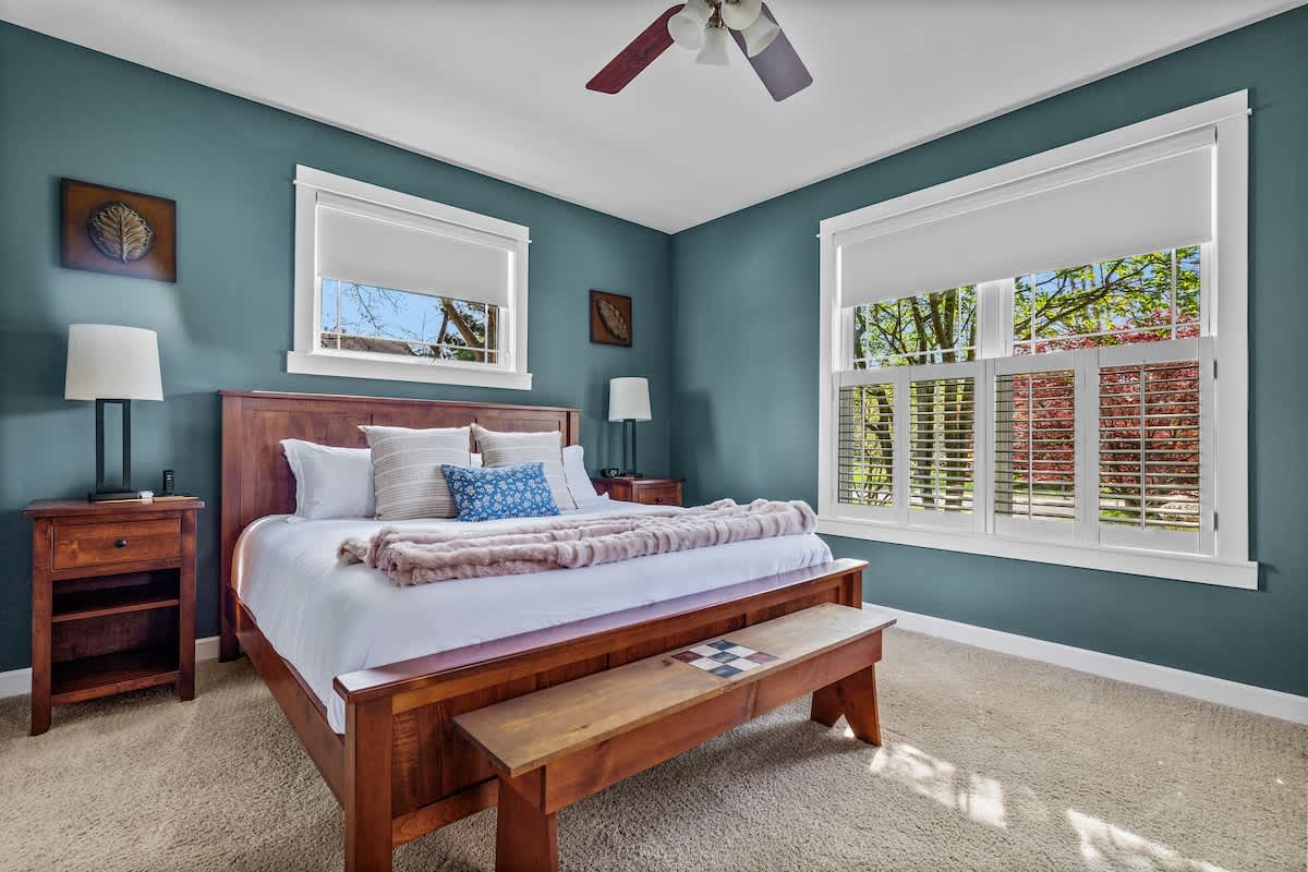 A spacious bedroom with teal walls features a wooden king-size bed with white bedding, flanked by matching nightstands and lamps. Large windows with white blinds and shutters overlook outdoor greenery, and a ceiling fan hangs overhead.