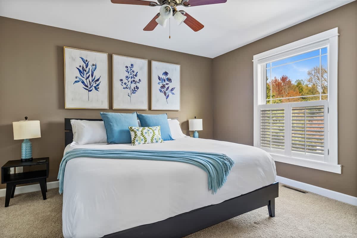 A cozy bedroom featuring a king-sized bed with white bedding and blue accents, warm brown walls, a ceiling fan, and a large window with white shutters overlooking autumn trees.