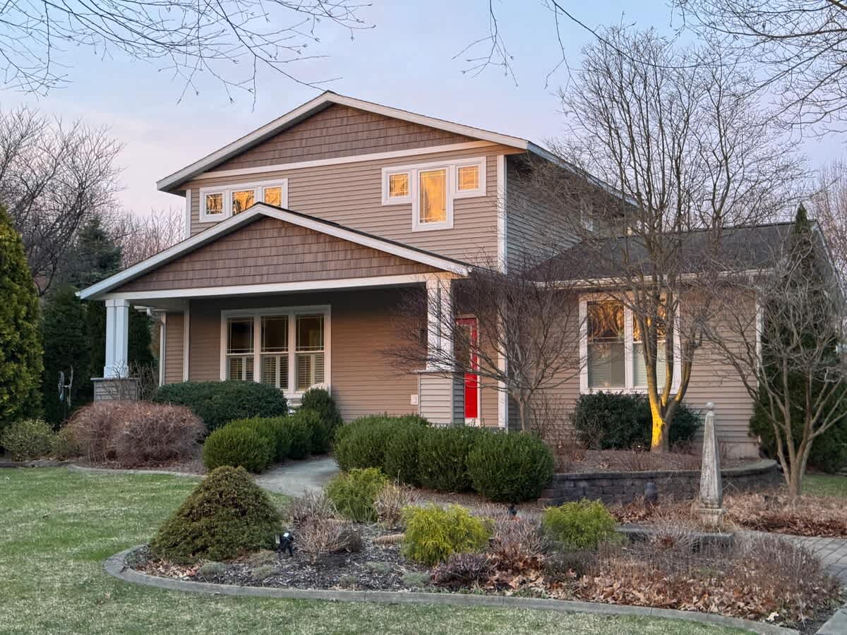 Front exterior view of a two-story house with light brown siding and darker shingle accents, featuring a covered porch and a red front door. The surrounding landscaping includes shrubs and bare trees, illuminated by soft evening light.