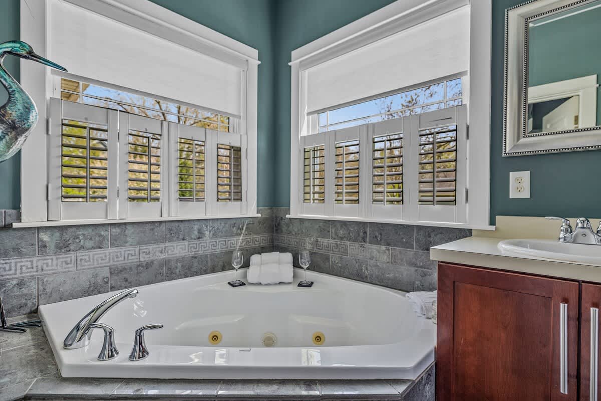 A close-up of a corner jetted bathtub with gray tile surround and white shutters on the windows above. Two wine glasses and folded towels are placed beside the tub, suggesting a relaxing atmosphere.
