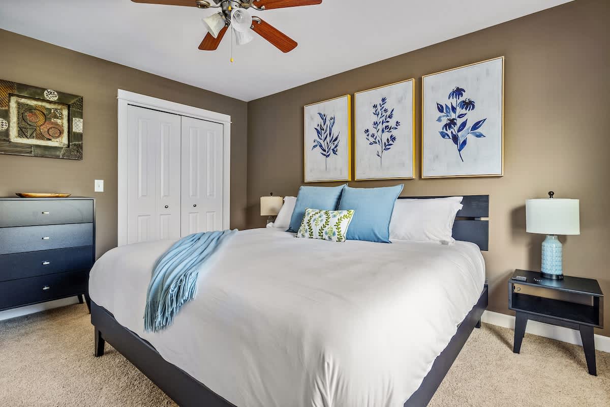 Wide shot of a bedroom with a king bed, white bedding, and blue accents, featuring warm brown walls, a dark dresser, a white bi-fold closet, and botanical artwork above the bed.