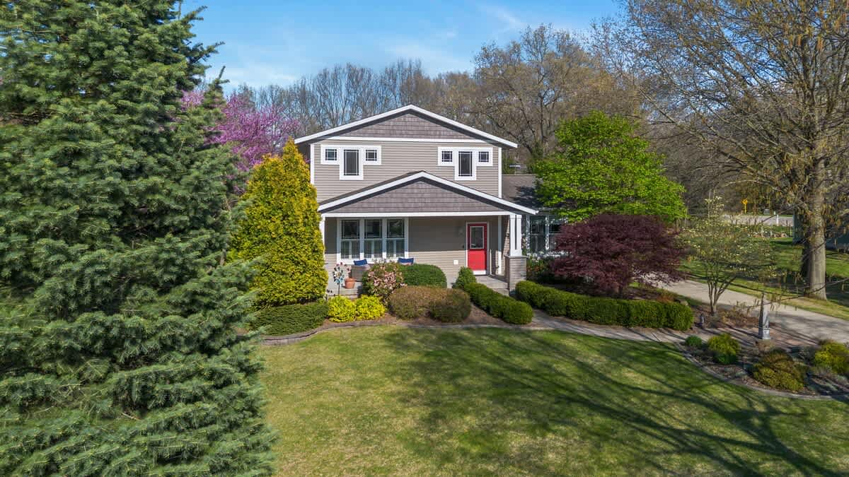 An inviting two-story house with gray siding, white trim, and a vibrant red front door, surrounded by expansive green lawns, mature trees, and colorful landscaping under a clear blue sky.