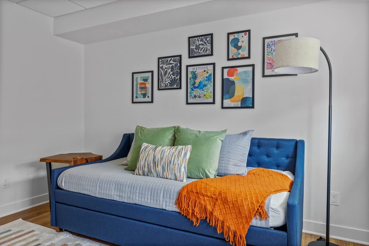 Cozy daybed in a vibrant blue upholstery with an orange throw blanket and an assortment of pillows, complemented by a gallery wall of framed art and a modern floor lamp.
