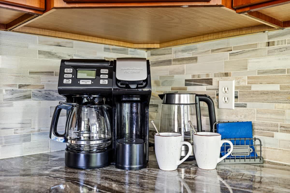 A close-up of a kitchen counter coffee station with a drip coffee maker, an electric kettle, and two white mugs, set against a modern tiled backsplash.