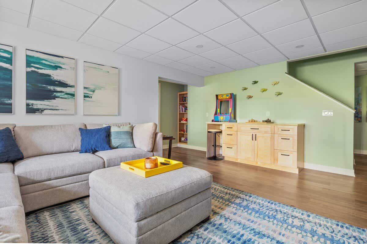 Spacious living area transitioning into a game zone with a classic arcade machine, built-in light wood cabinetry, and a bookshelf, all beside a comfortable sectional sofa.