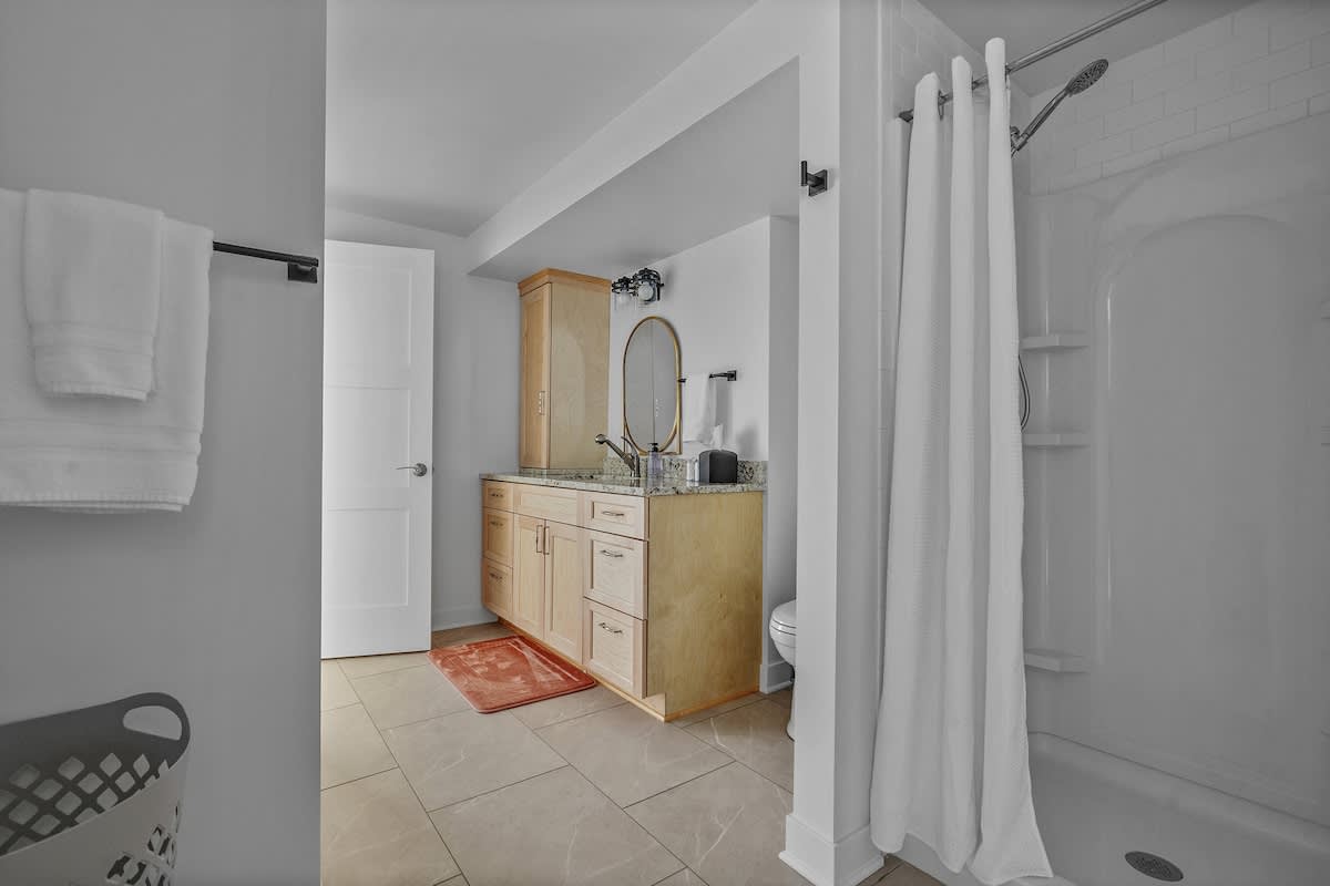 A bright bathroom with a light wood vanity, granite countertop, oval mirror, and a white shower with a curtain, featuring light gray walls and tiled flooring.