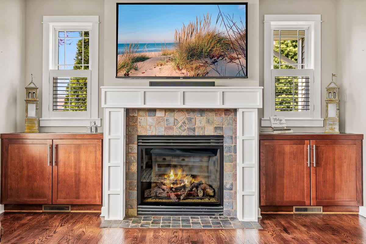 A cozy living room fireplace with a tiled surround and a white mantel, topped by a large flat-screen TV displaying a beach scene, flanked by dark wood built-in cabinets.