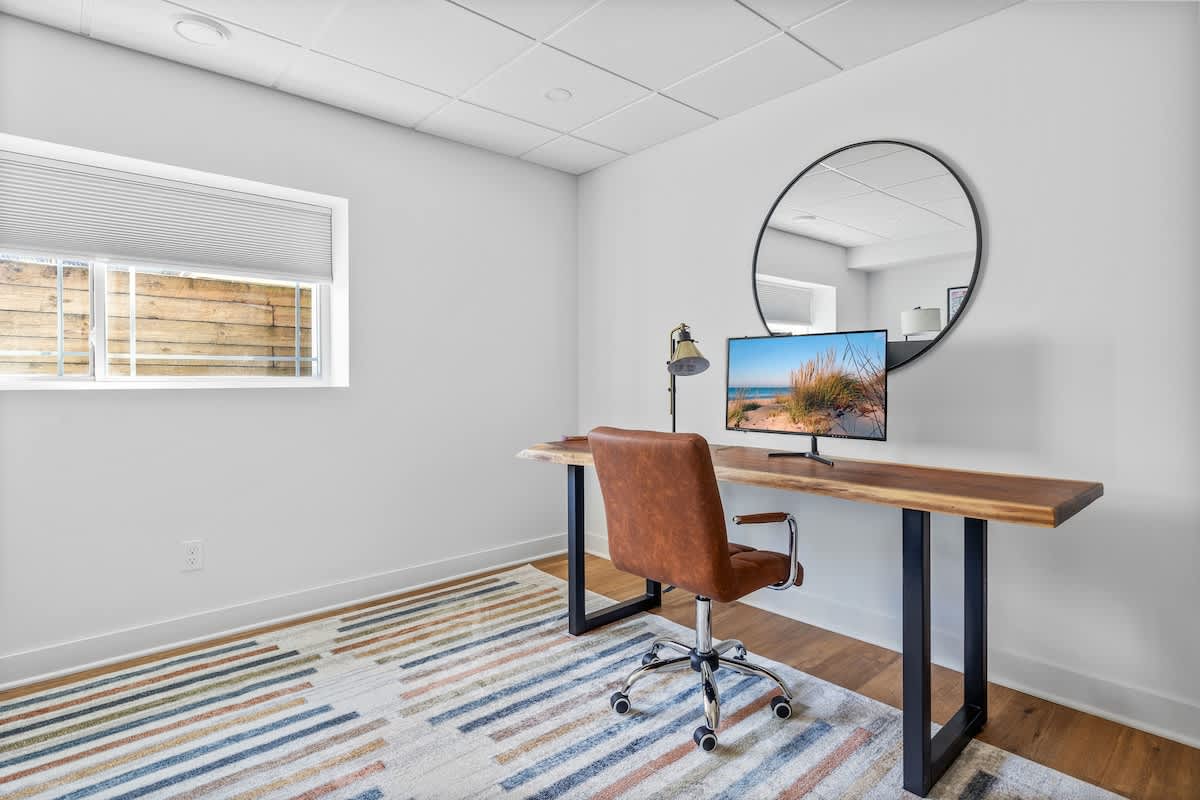 Modern home office with a live-edge wooden desk, brown leather office chair, and a large round mirror, set on a colorful geometric rug. A window with blinds offers natural light.