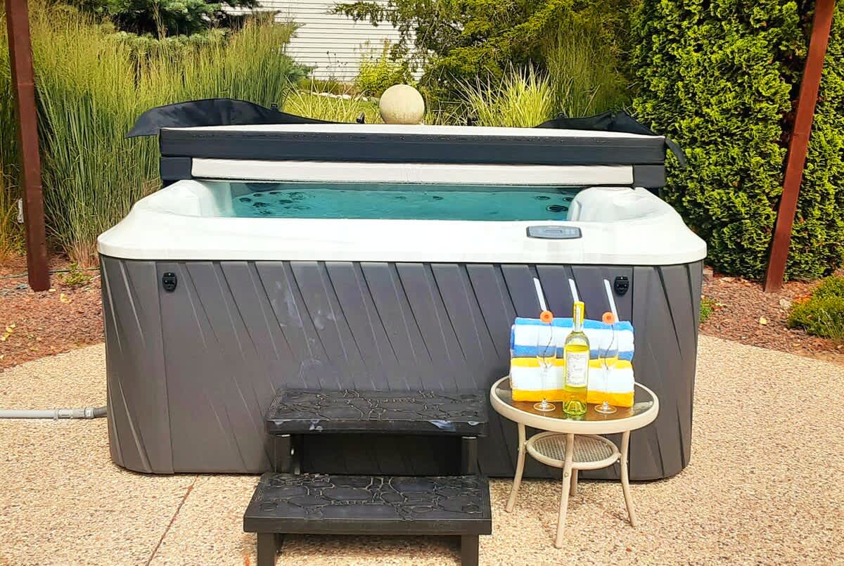 Close-up view of a hot tub with its cover partially open, showing clear water. A small side table holds two glasses with drinks and striped towels, set on a textured concrete patio surrounded by green foliage.