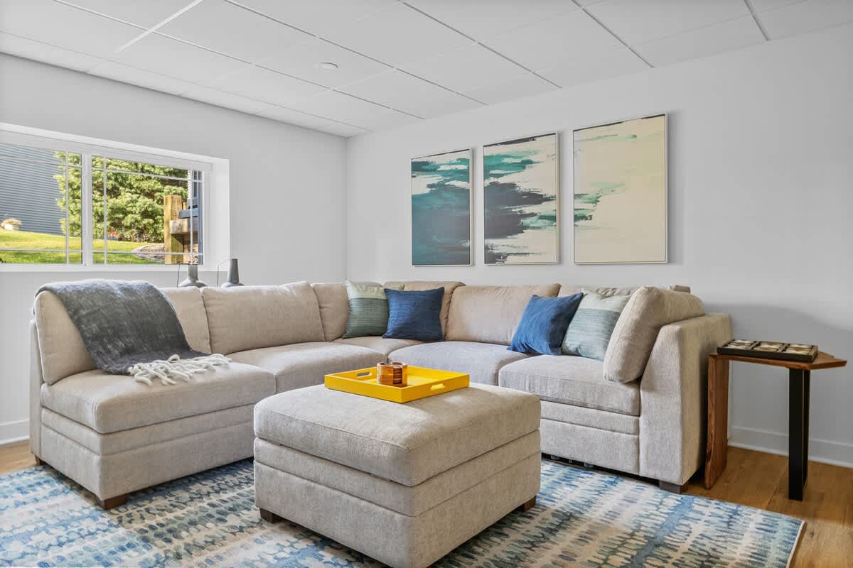 Inviting living room with a large light-colored sectional sofa, a matching ottoman with a yellow tray, and a trio of abstract blue and white artworks above, all on a patterned rug.