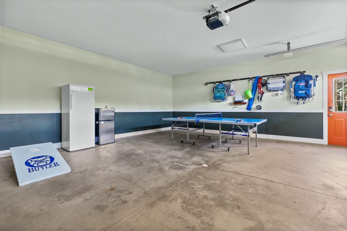 Spacious garage converted into a recreation area with a blue ping pong table, a cornhole game, a full-size refrigerator, and wall-mounted storage for outdoor gear.