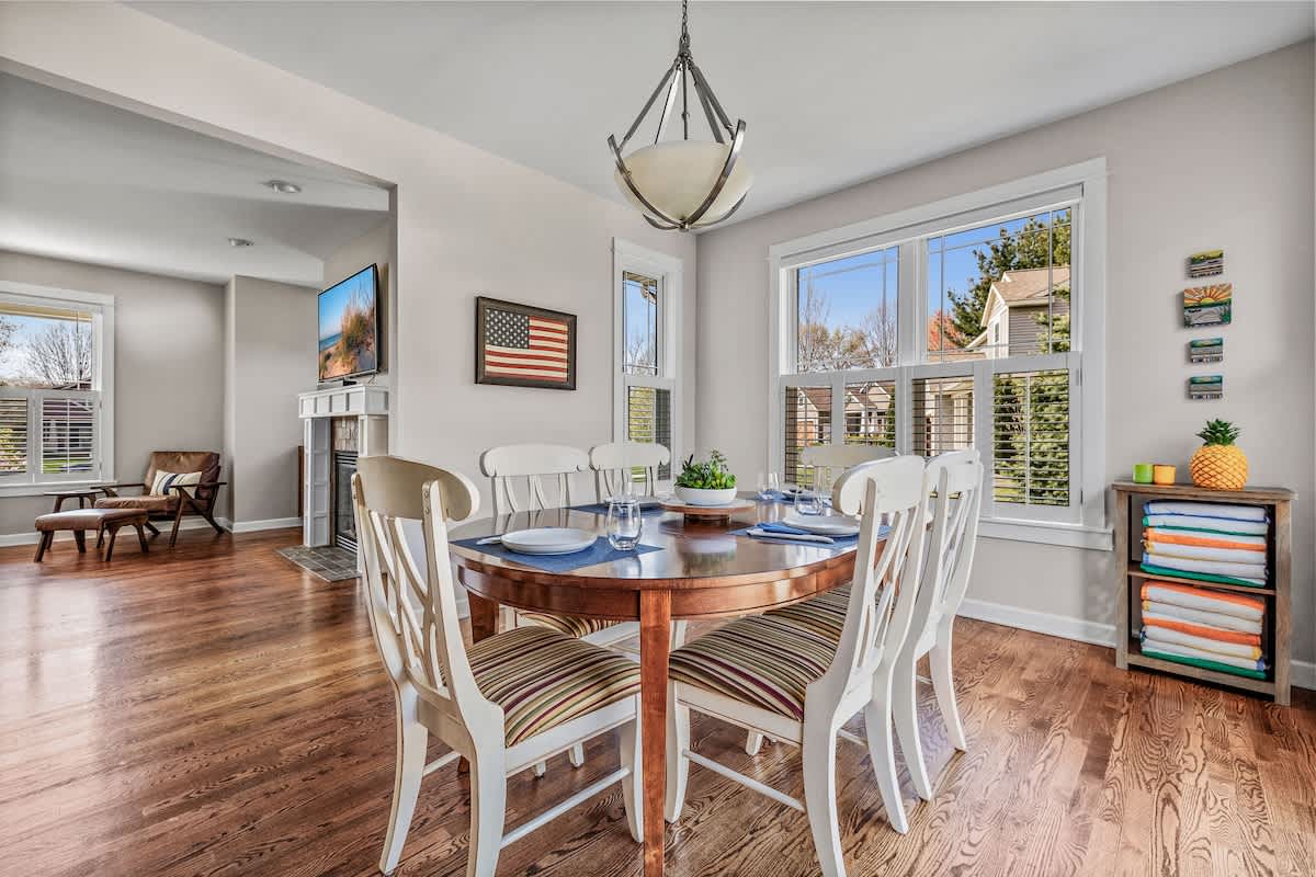 A bright dining room with a round wooden table set with place settings and surrounded by white chairs, featuring large windows with white blinds and hardwood floors.