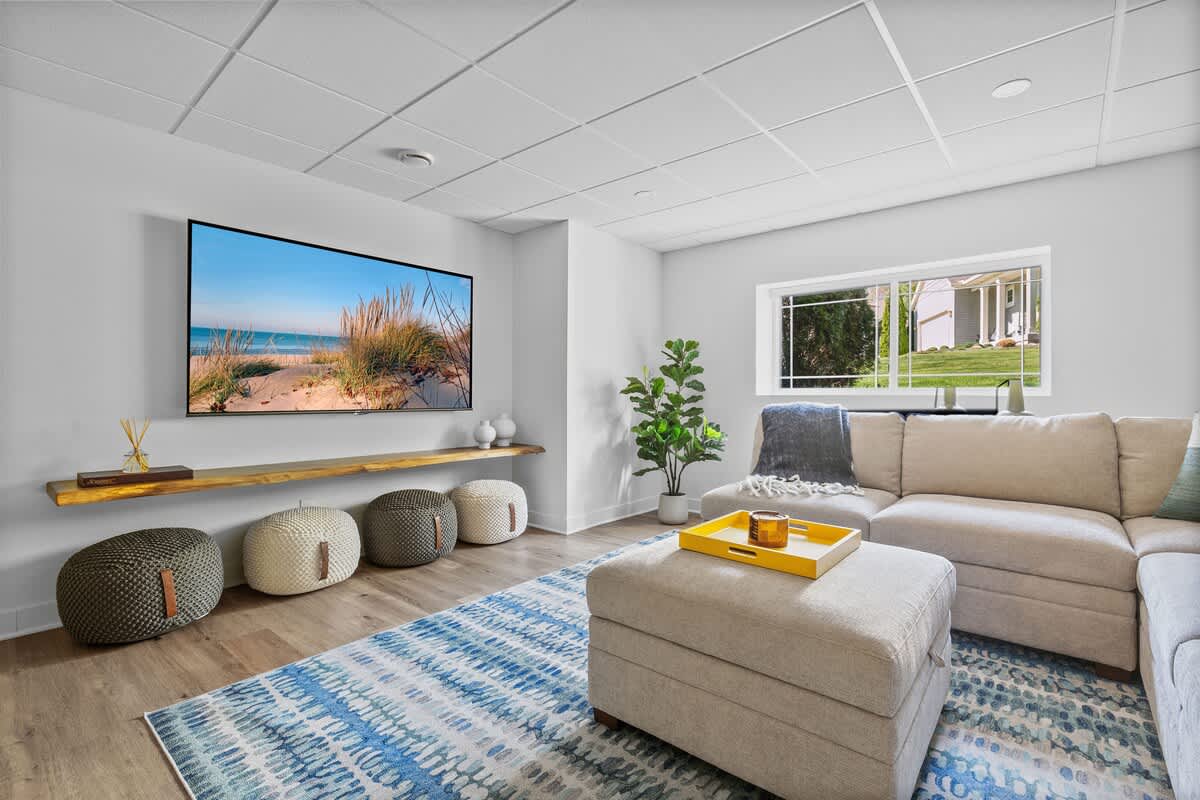 Bright living room featuring a large light-colored sectional sofa, a wall-mounted TV displaying a beach scene, and a blue and white patterned rug, with natural light from a large window.