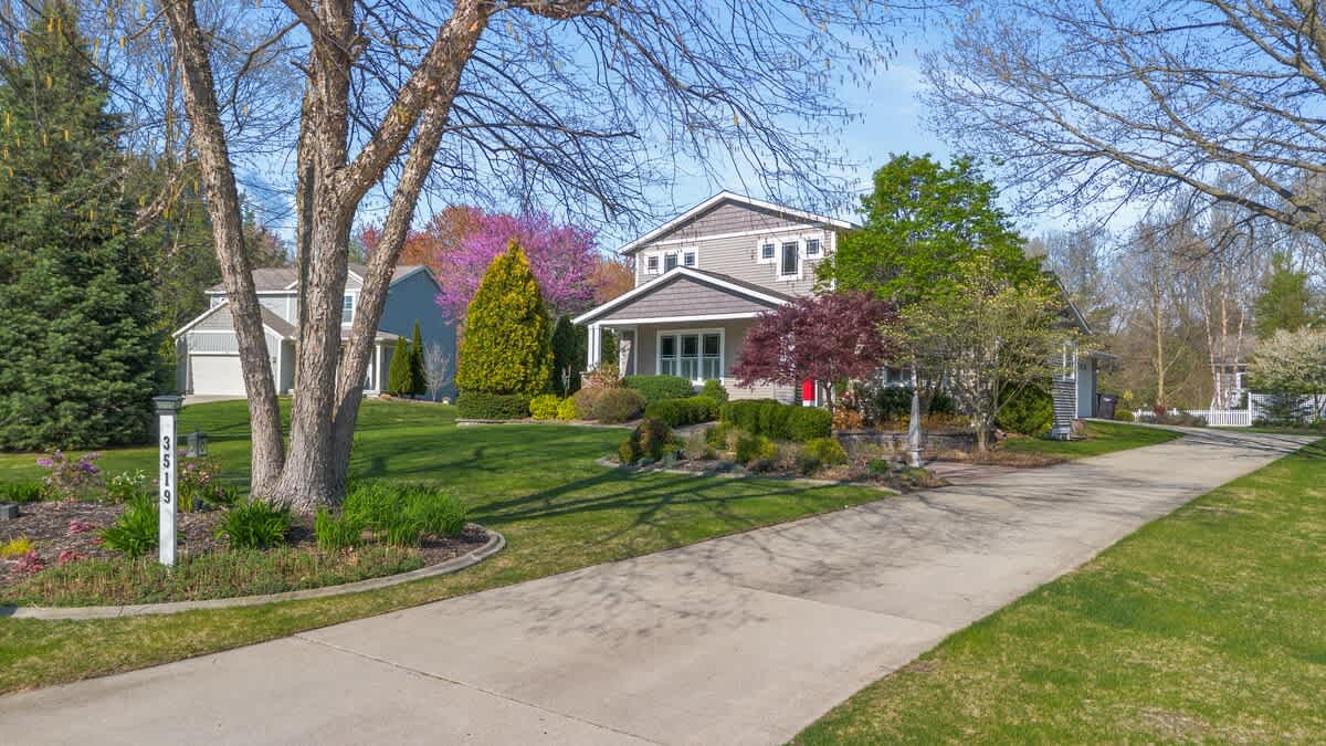 Wide exterior view of the house from the driveway, showcasing its light brown siding and varied rooflines, surrounded by a well-maintained lawn and mature trees with spring foliage. A paved driveway leads up to the house.
