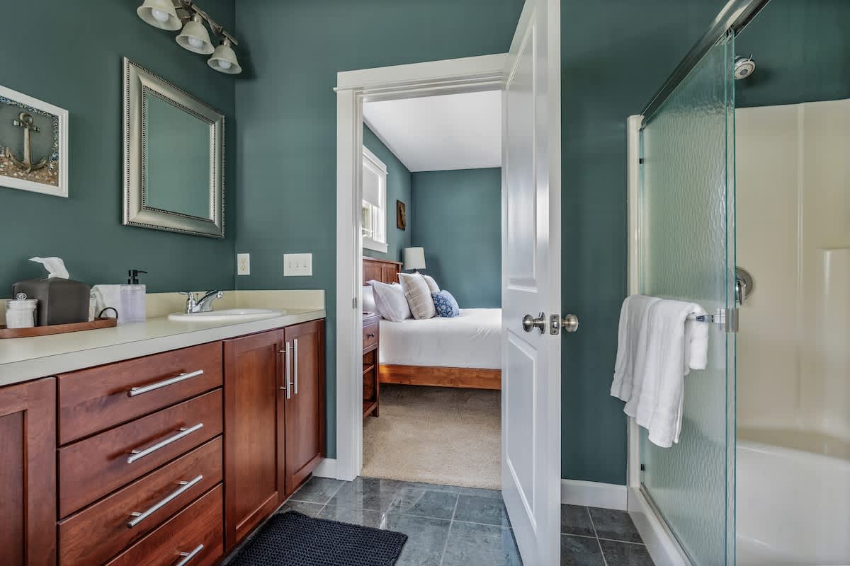 A bathroom with teal walls features a single vanity with a wooden cabinet and white countertop, and a shower/tub combo with a glass door. An open doorway provides a glimpse into the adjacent bedroom.
