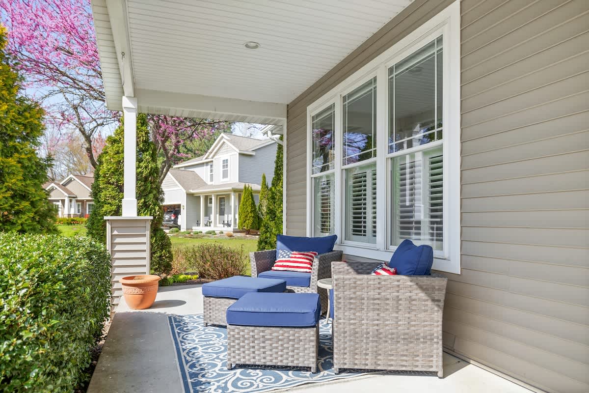 Inviting covered front porch with comfortable wicker-style seating, including a sofa and ottoman, adorned with blue cushions and patriotic pillows. A patterned blue rug defines the seating area, overlooking a landscaped front yard.