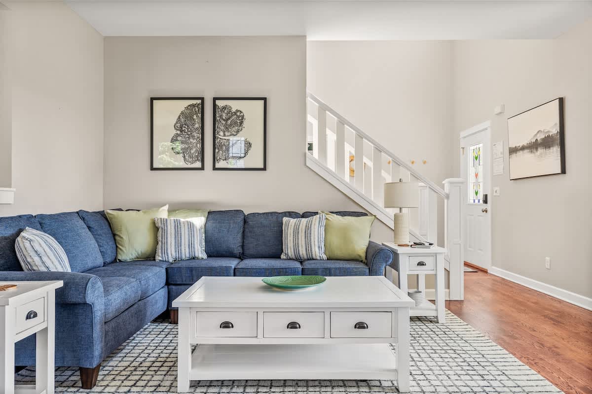 A bright living room with a large blue sectional sofa, white end tables, and a white coffee table on a patterned rug, with a white staircase leading to the upper level.