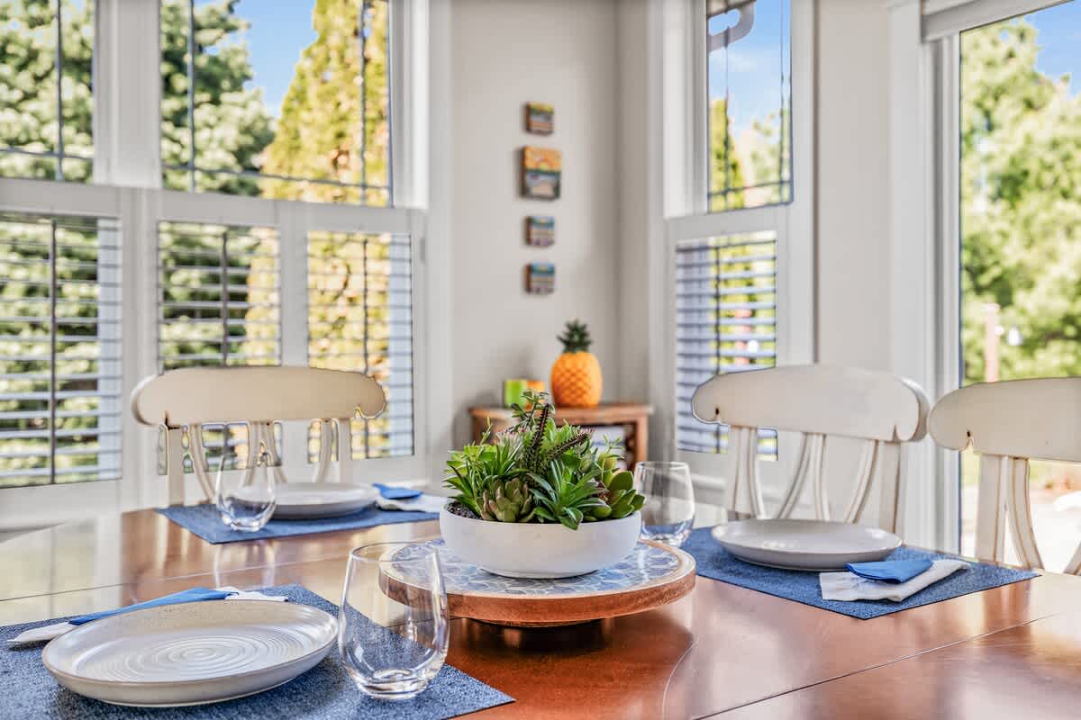 A bright dining area features a polished wooden table set with blue placemats, white plates, and clear glasses, centered by a succulent arrangement on a wooden riser. Large windows with white frames offer views of lush greenery outside.