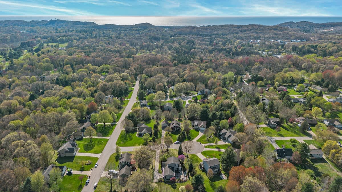 An aerial view of a residential neighborhood with many houses nestled among abundant green trees, leading towards a large body of water under a blue sky with scattered clouds.