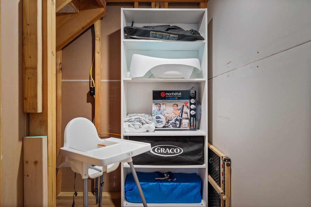 A dedicated storage area or utility space featuring shelves stocked with various baby and toddler essentials, including a white high chair, a portable potty, a Pack 'n Play, and a baby gate, set against unfinished plywood and drywall walls.