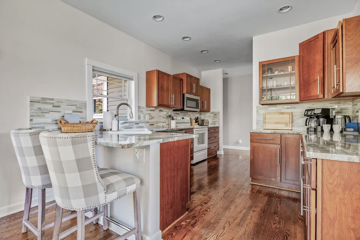 A bright kitchen featuring a breakfast bar with two plaid upholstered stools, dark wood cabinets, light countertops, and a window overlooking the exterior, with hardwood floors throughout.