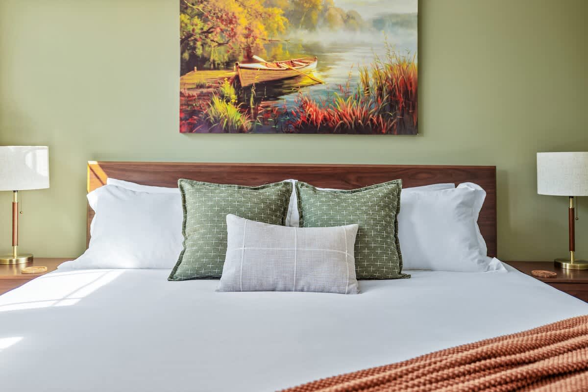 A close-up of a king-size bed with a modern wooden headboard, white linens, and decorative green and gray pillows. A vibrant landscape painting depicting a boat on a misty lake hangs above the bed.
