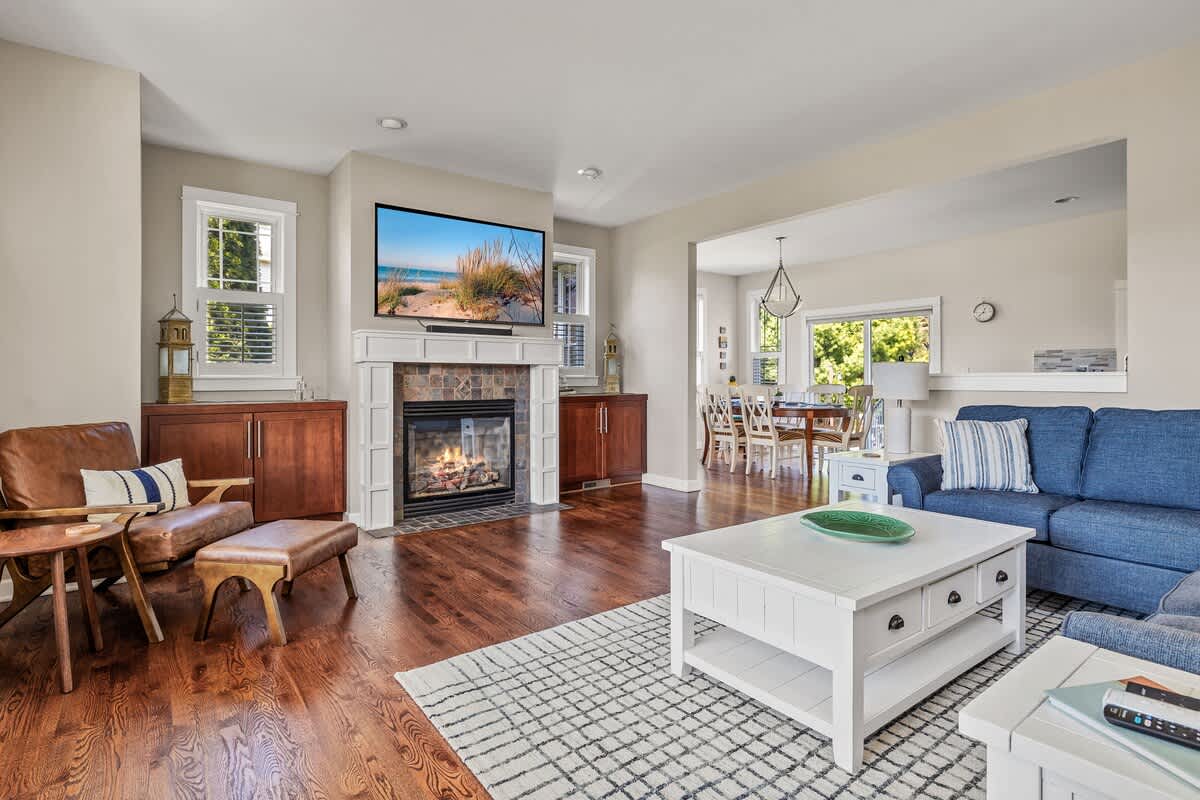 Cozy living room featuring a stone-faced fireplace with a TV mounted above, flanked by built-in wooden cabinets. A blue sofa and a brown leather armchair are arranged around a white coffee table on a patterned rug, with hardwood floors throughout.