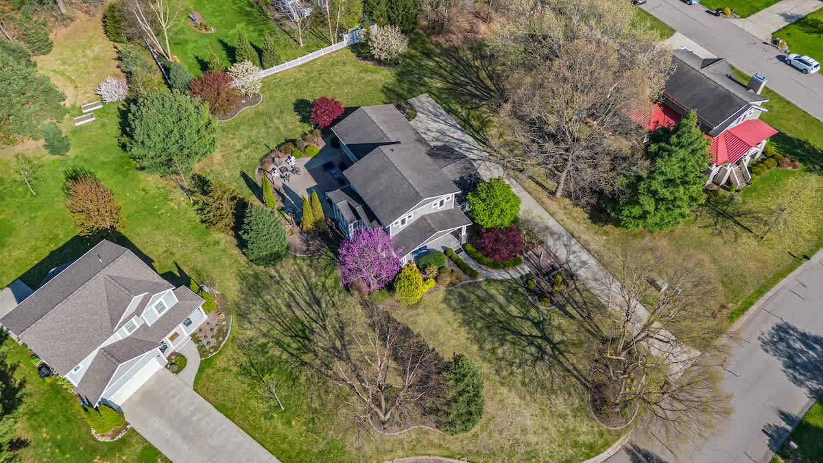 An expansive aerial view showcasing a large residential property with a gray house, extensive green lawns, mature trees, and colorful spring foliage, nestled within a well-maintained suburban neighborhood.