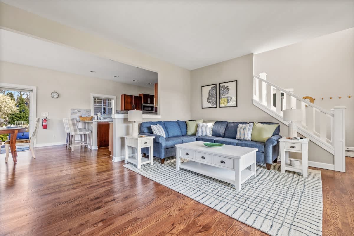 A spacious living room featuring a large blue sectional sofa with throw pillows, a white coffee table, and a staircase with white railings leading upstairs, all on rich hardwood floors.