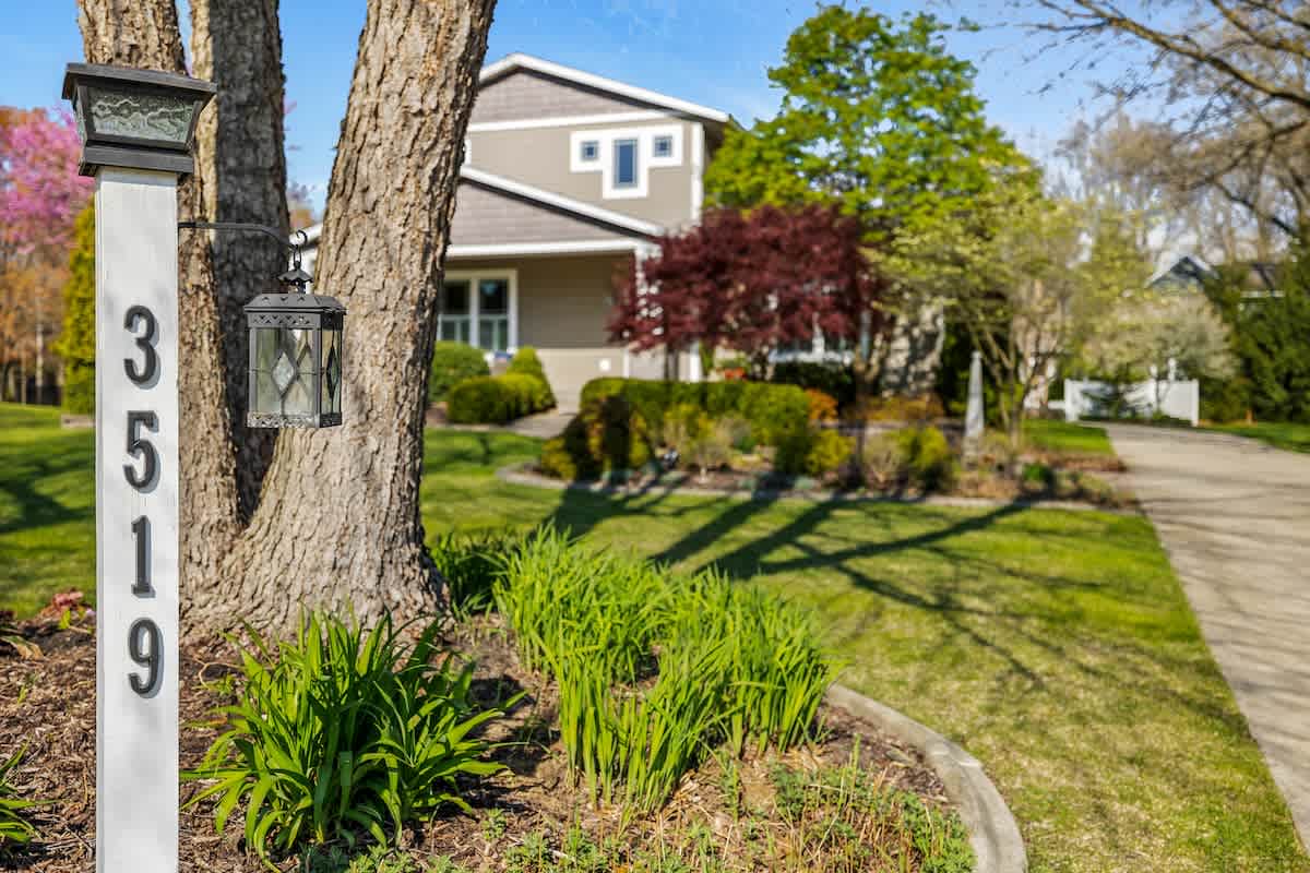 A charming white house number post with a decorative lantern, displaying the numbers '3519', stands beside a mature tree and lush green landscaping, with a modern gray house visible in the background under a bright blue sky.