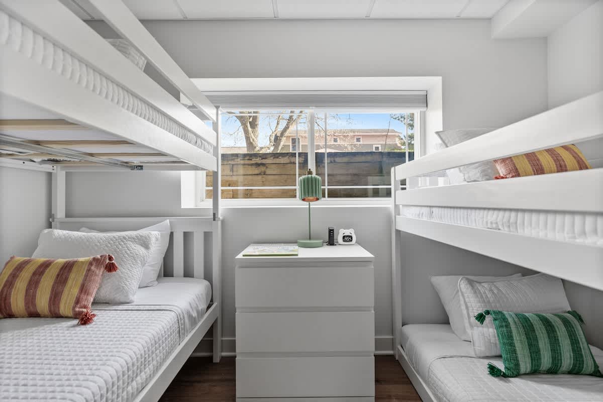 A bright children's bedroom featuring two sets of white bunk beds with white bedding and colorful striped pillows, a white dresser, and a window overlooking a fence.