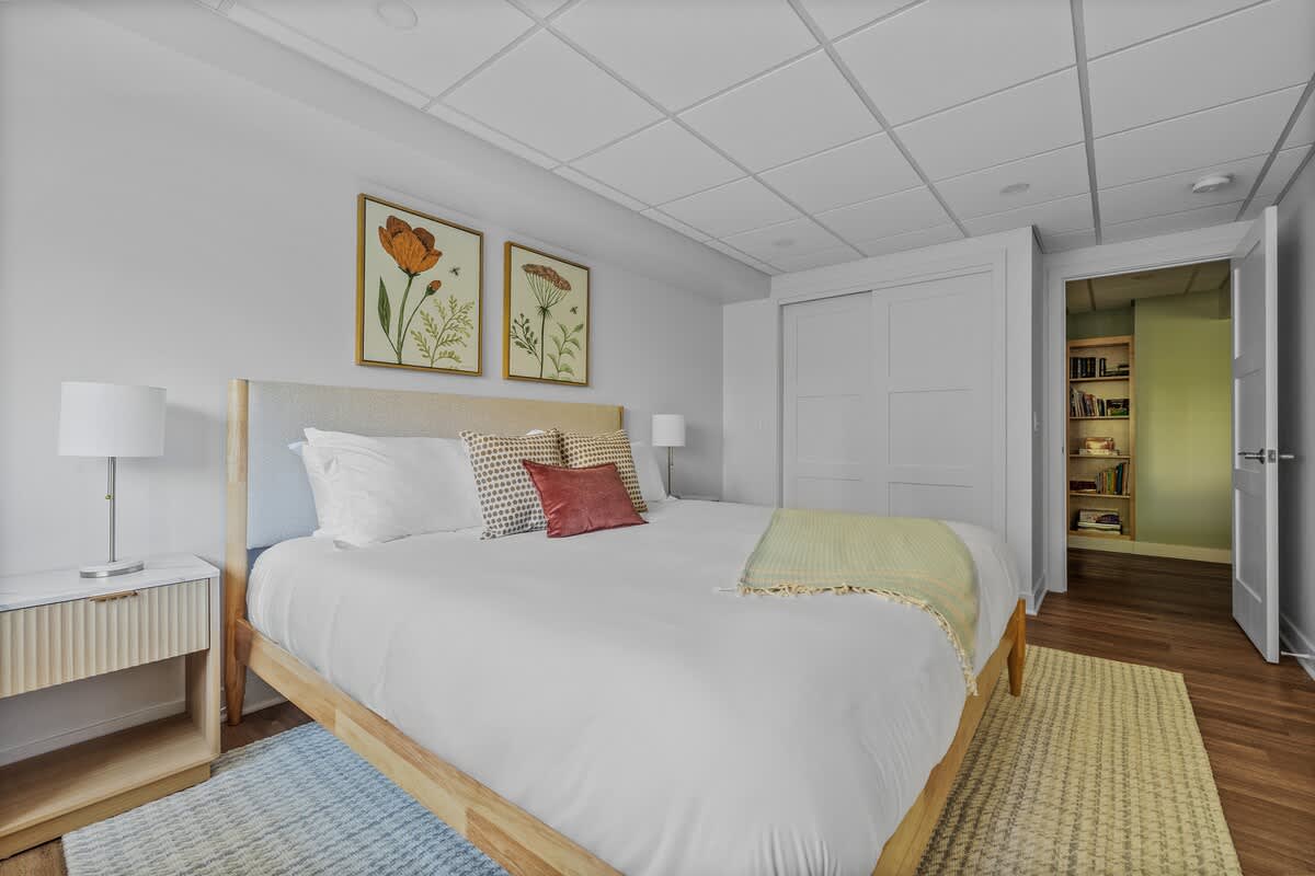 A spacious bedroom with a king bed, light wood frame, and white bedding, featuring white walls, a large closet with sliding doors, and an open doorway leading to another room.