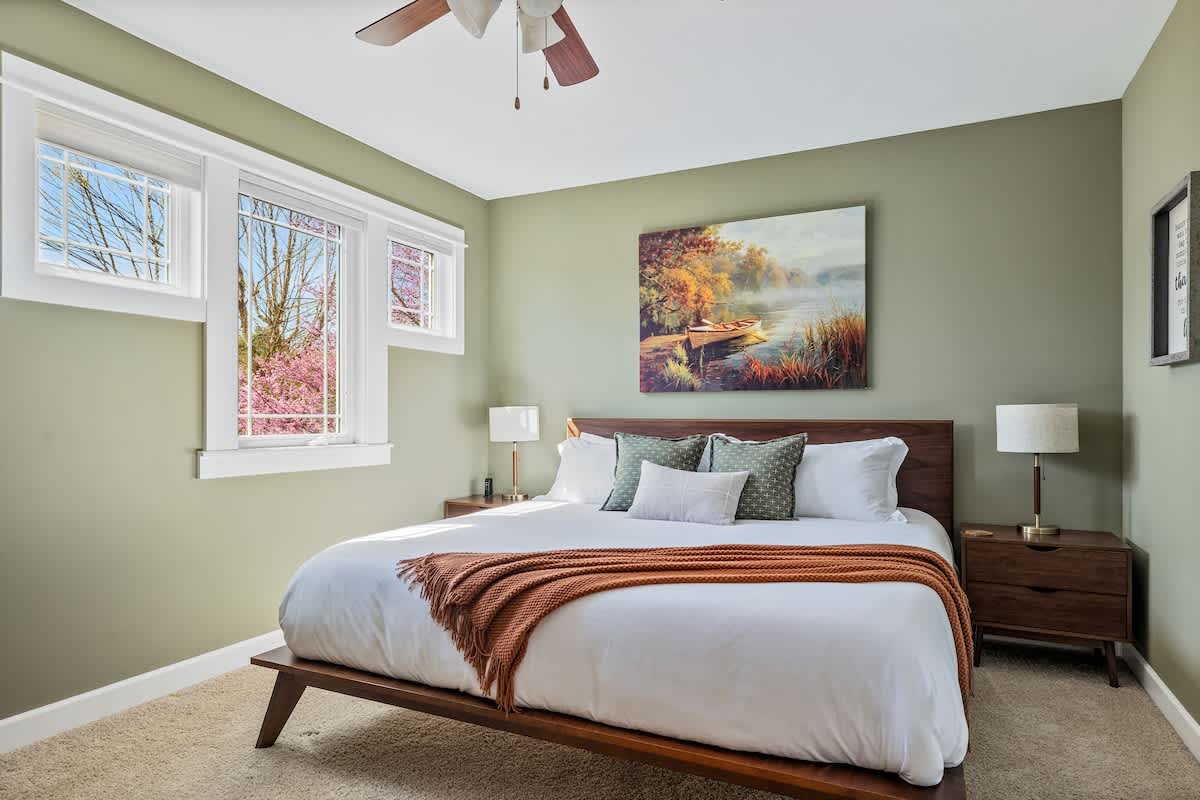 A serene bedroom with light green walls features a modern wooden bed frame with a white mattress and an orange throw blanket. A large landscape painting hangs above the headboard, and two windows offer views of trees.