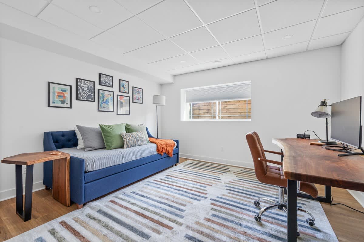 A versatile room featuring a blue daybed with colorful pillows, a large wooden desk with a computer monitor, a striped area rug, and a gallery wall of framed art.