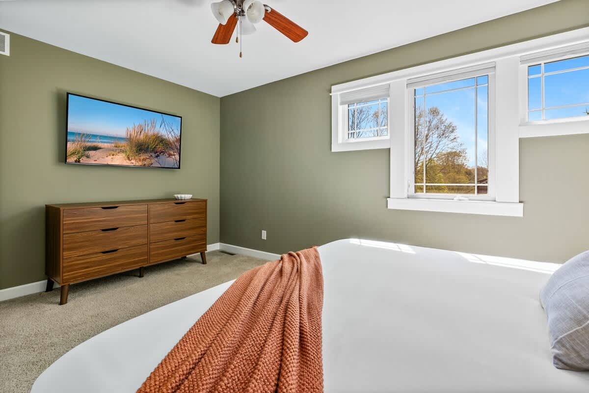A bedroom with light green walls features a wall-mounted flat-screen TV above a modern wooden dresser with multiple drawers. A king-size bed with white bedding and an orange throw is partially visible, and windows offer natural light.