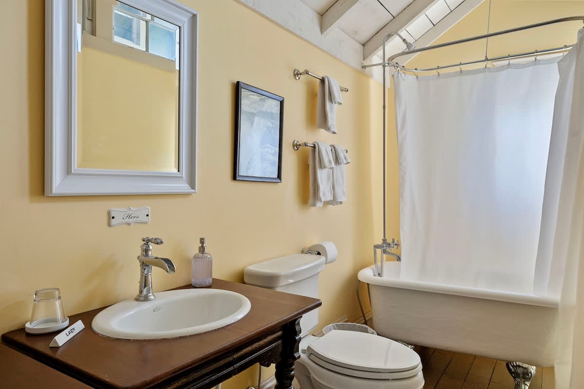 Bathroom with light yellow walls, a classic clawfoot tub with a shower curtain, and a wooden vanity with a white basin sink. A large mirror hangs above the sink.