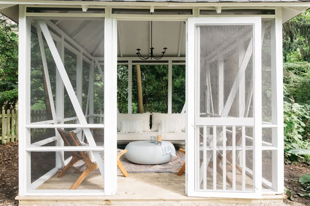 White screened-in gazebo with a comfortable daybed, a round white coffee table, and two wooden lounge chairs, surrounded by lush greenery. A small chandelier hangs from the ceiling.
