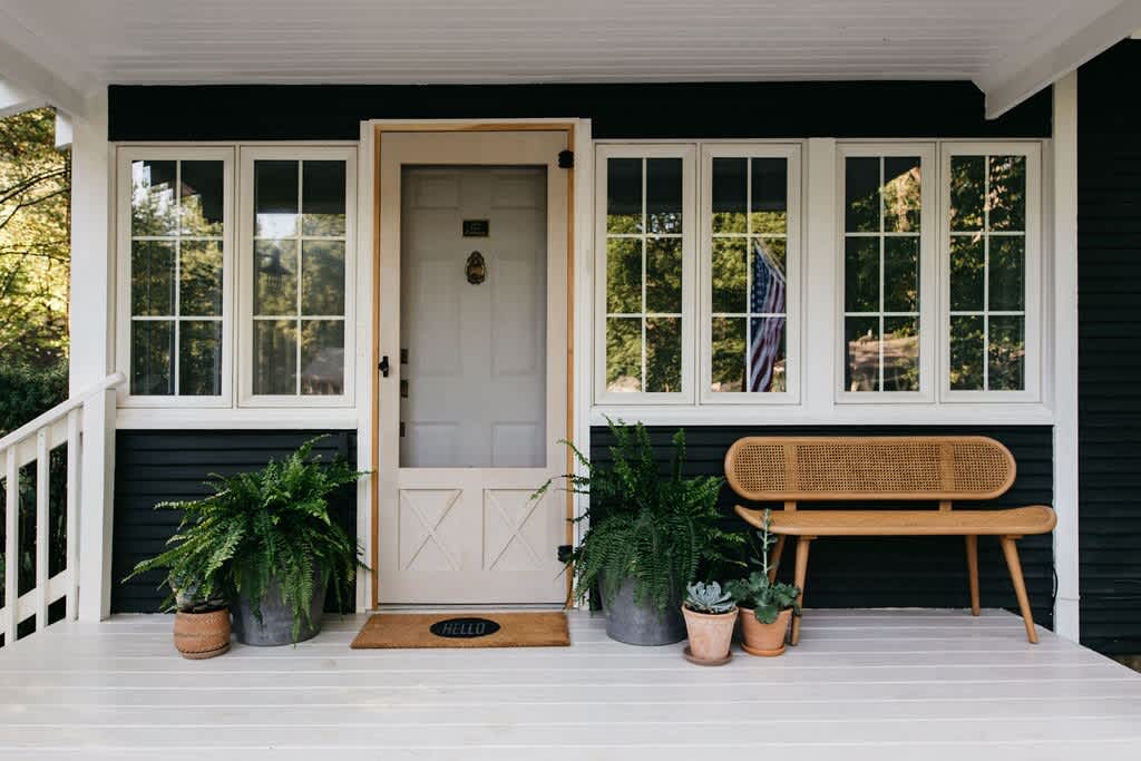 Welcoming front porch with a light-colored door, a 'HELLO' mat, a wooden bench with cane detailing, and several potted plants, all set against a dark house with white-trimmed windows.