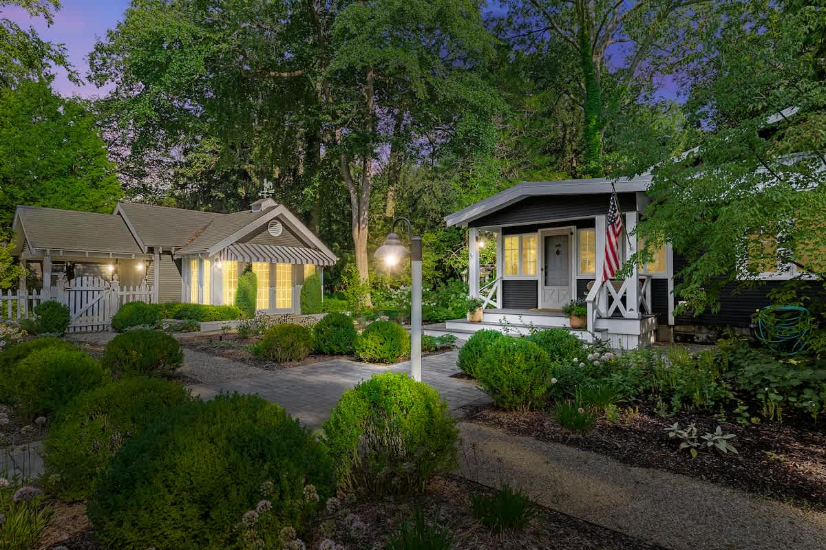 An evening exterior view of multiple charming cottage-style buildings with dark siding and white trim, illuminated by outdoor lights. A paved pathway leads through manicured bushes and gardens.